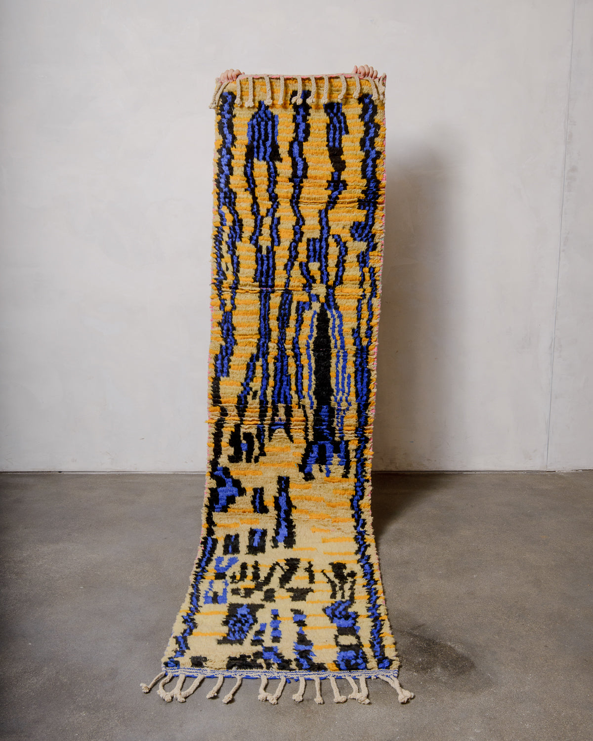 Berberlin vintage Berber runner rug made of colourful wool, being held up in front of a white wall and concrete floor.